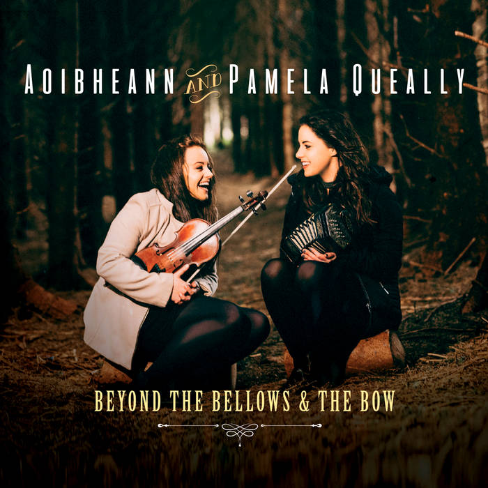 Two women sit on logs in a forest. One wears a white jacket and is laughing while holding a fiddle. The other smiles at the first women, while she holds a concertina.