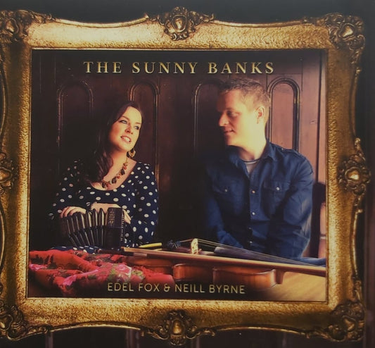 A square framed album cover or print featuring two musicians seated indoors, with the text 'THE SUNNY BANKS' at the top and 'EDEL FOX & NEILL BYRNE' at the bottom. The artwork is enclosed in an ornate gold frame against a dark background.